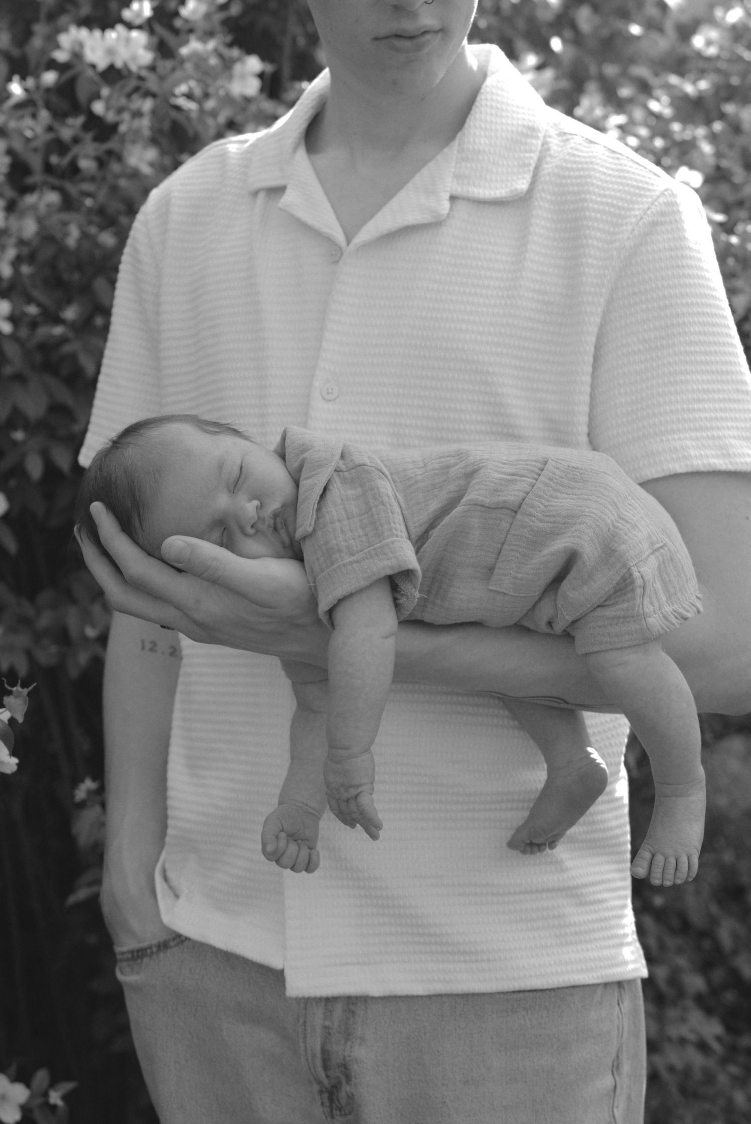 Parent holding sleeping newborn baby while wearing white polo shirt outdoors.