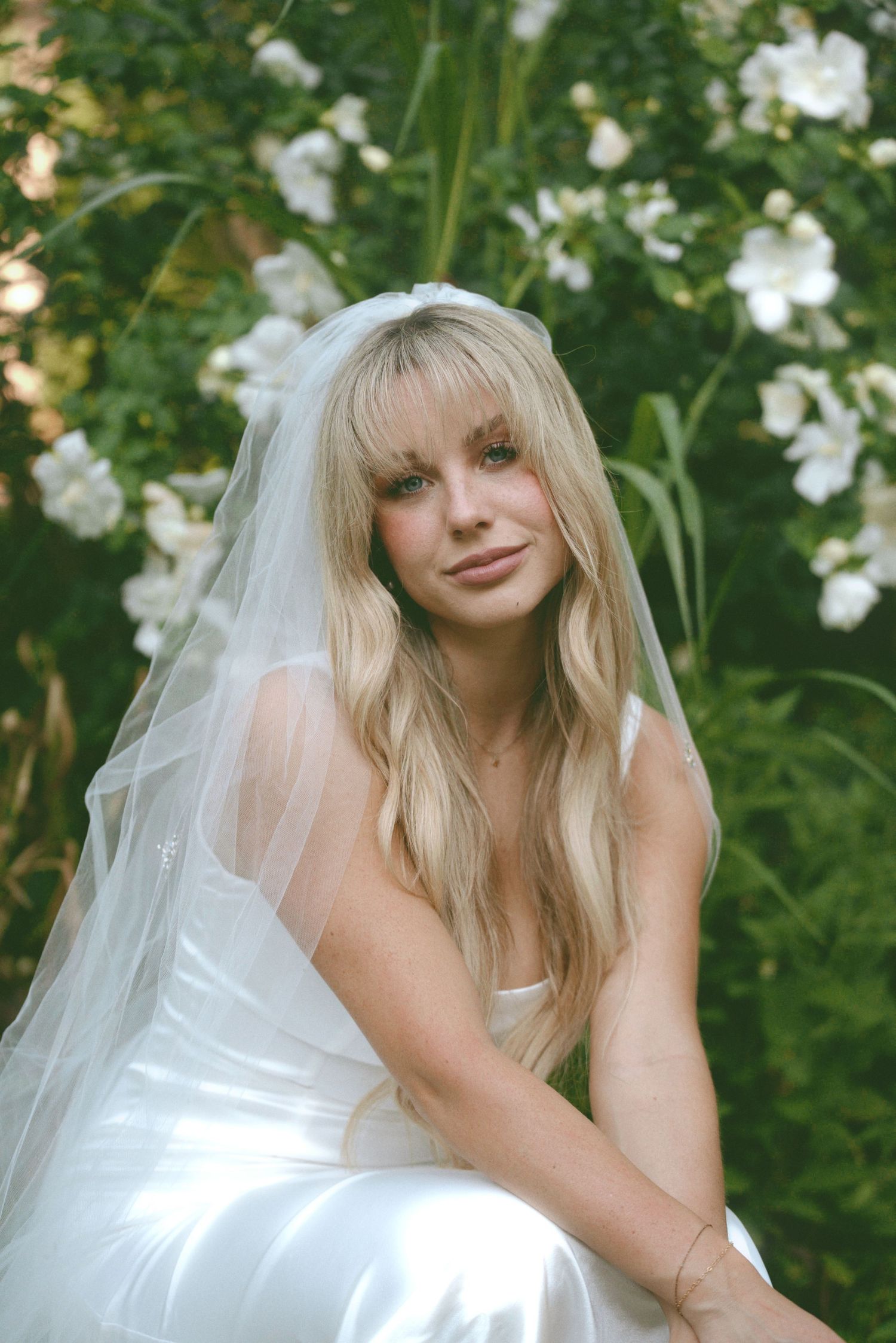 A bride in a white wedding dress and long veil poses in a garden with white flowers and green foliage in the background.