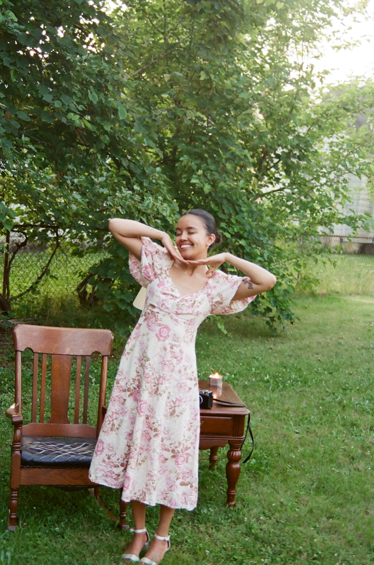 A candid summer moment in a garden with someone stretching while wearing a pink floral midi dress beside vintage furniture.