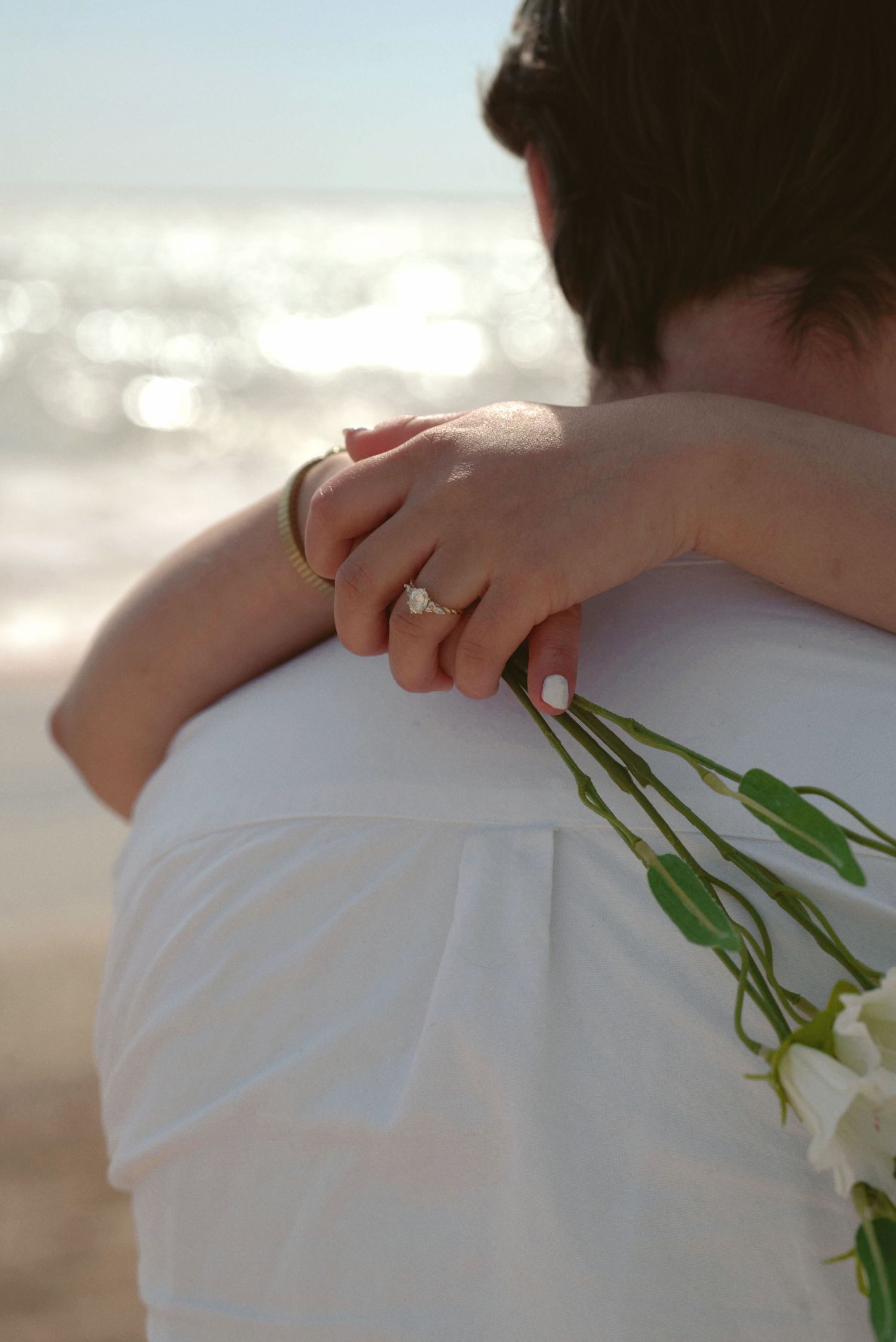 Newlywed couple embracing at sunset with green flowers visible against white wedding attire.