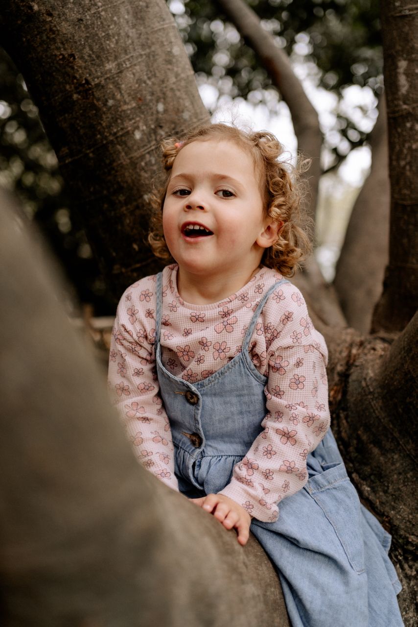 3 year old girl climbing a tree and looking at the camera with a cheeky smile at an outdoor family photoshoot in Hampshire