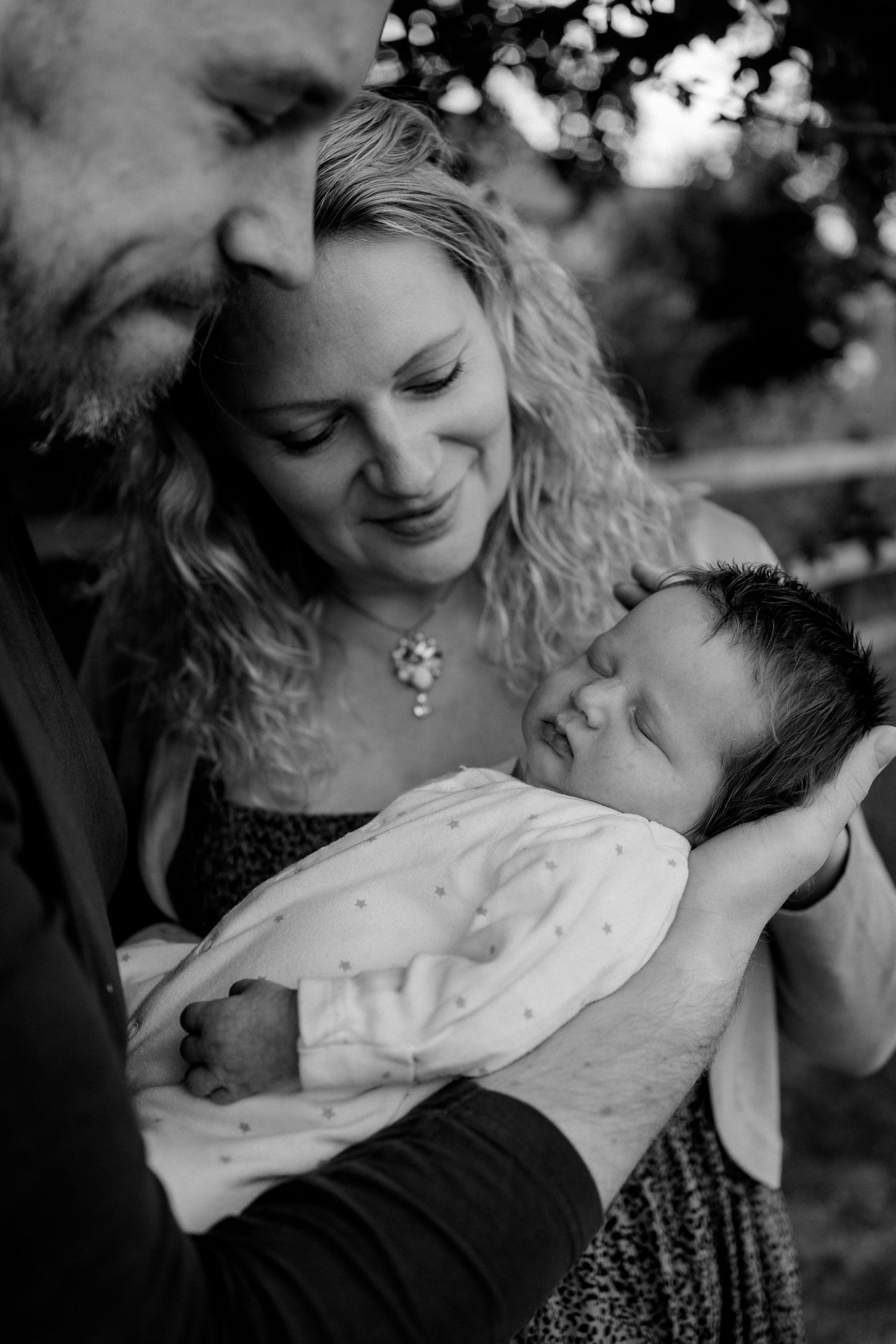 Dad holding a newborn baby while the mum looks at the baby with loving eyes and a gentle smile in their garden in Surrey
