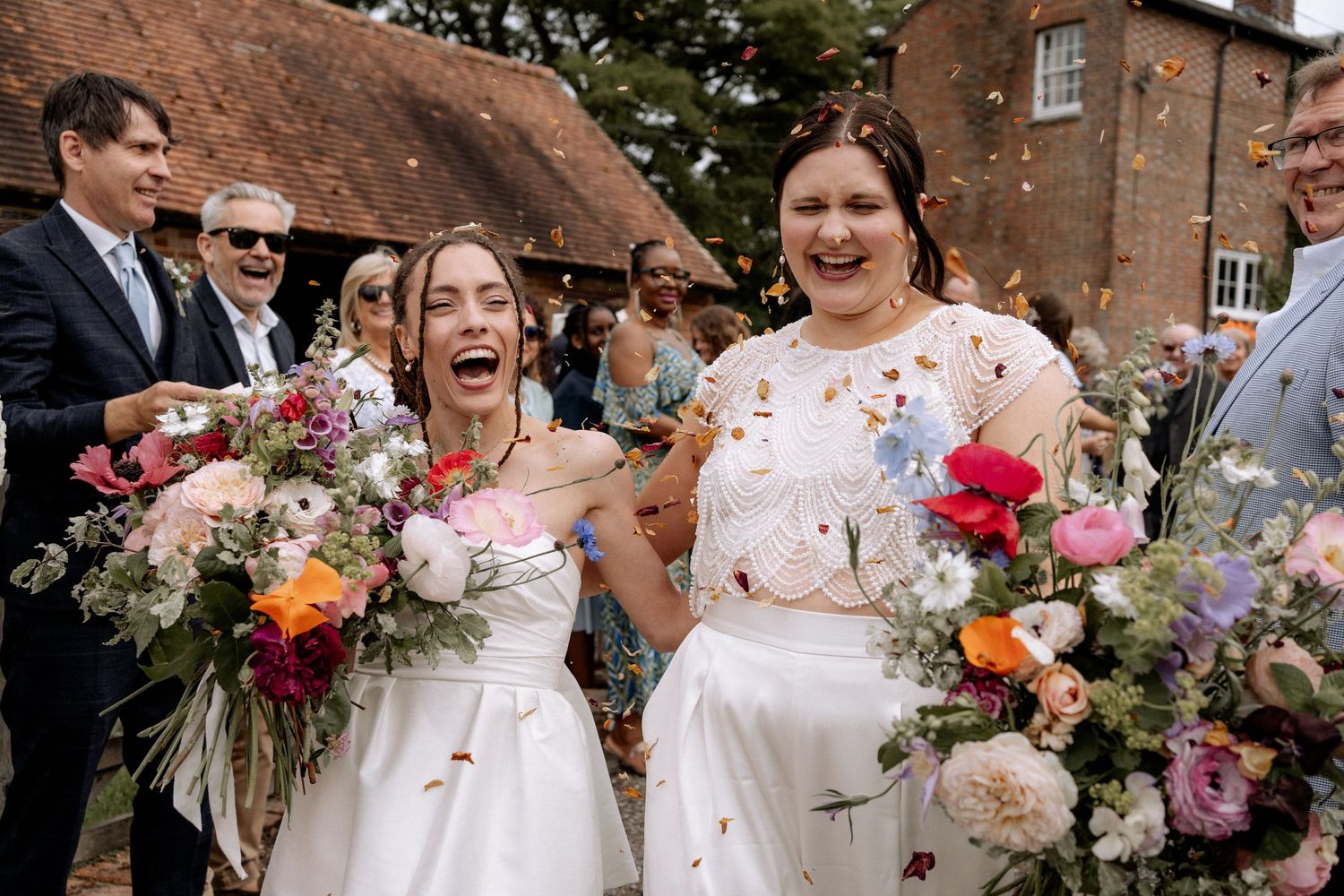 Two brides during their confetti throw, laughing with joy, at their wedding at the Secret Barn in Steyning, Sussex