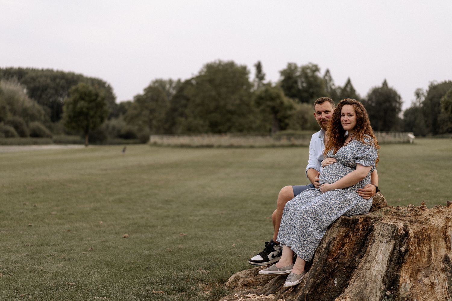 Man and woman sitting on a tree stump in a park, male partner hugging his pregnant partner who is holding her bump in Oxfordshire