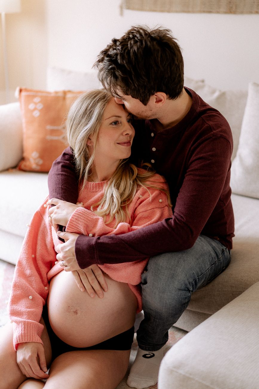 Male and female cuddling on the sofa. Male partner sits behind the pregnant woman, hand touching her pregnant belly at their home in Surrey