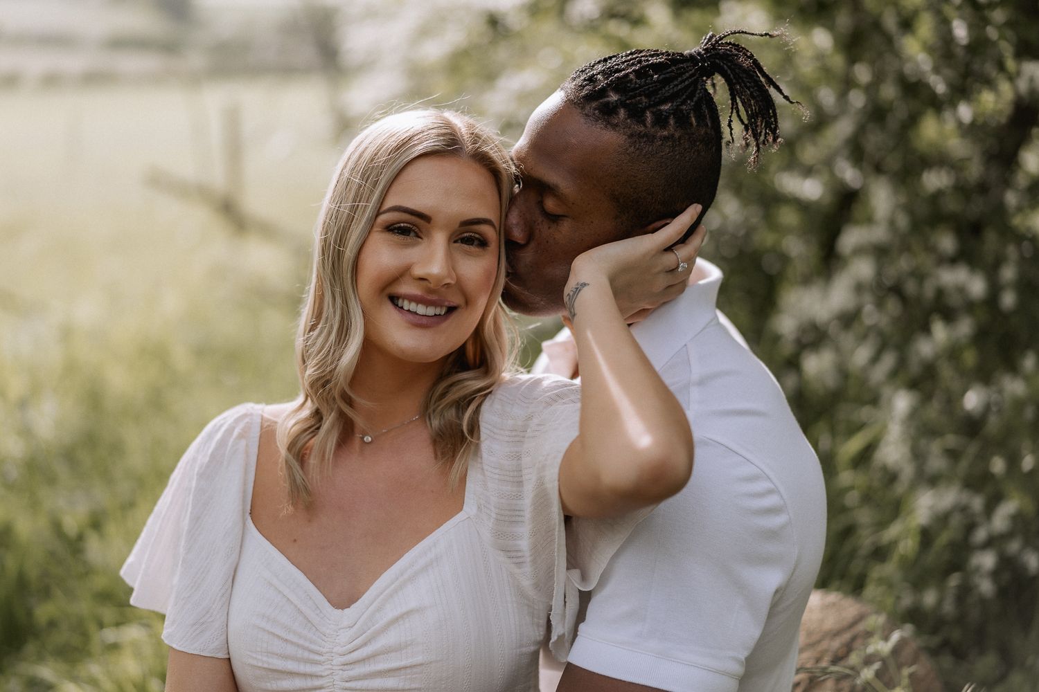 Male partner kissing his female partner on the side of the forehead while she smiles directly at the camera at an outdoor engagement shoot