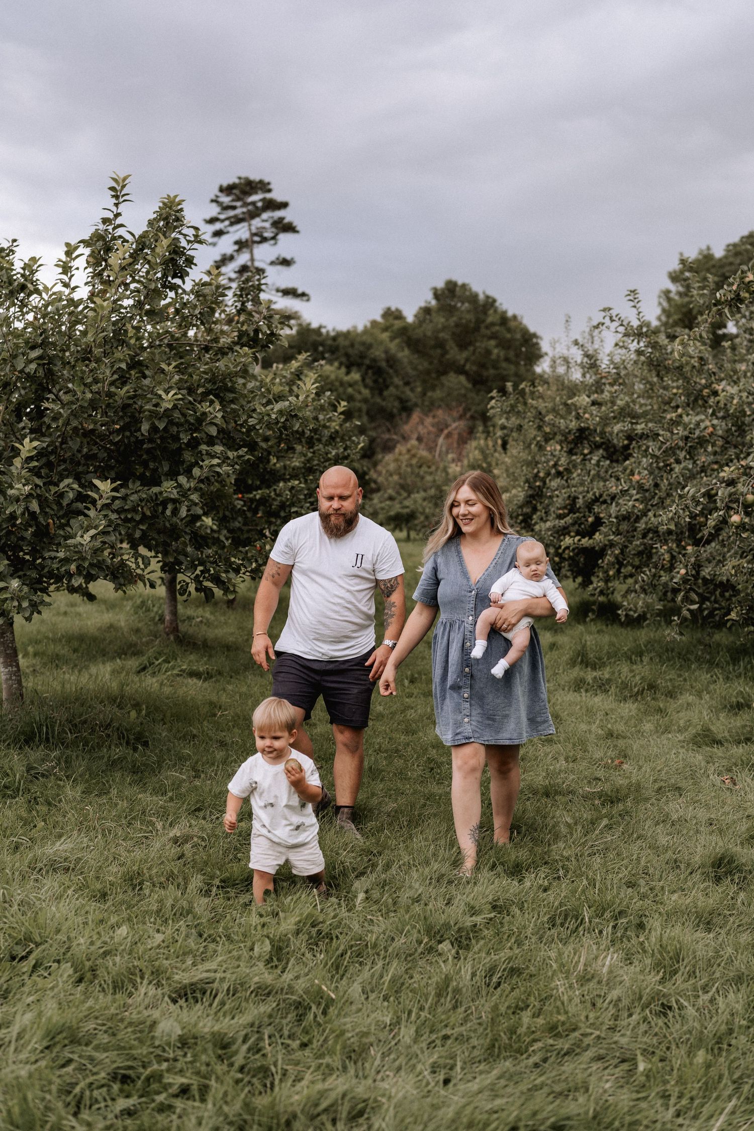 Family with dad, mum and toddler walking through an orchard in summer, coordinated outfits. Mum holds the baby. Outdoor photos in Surrey