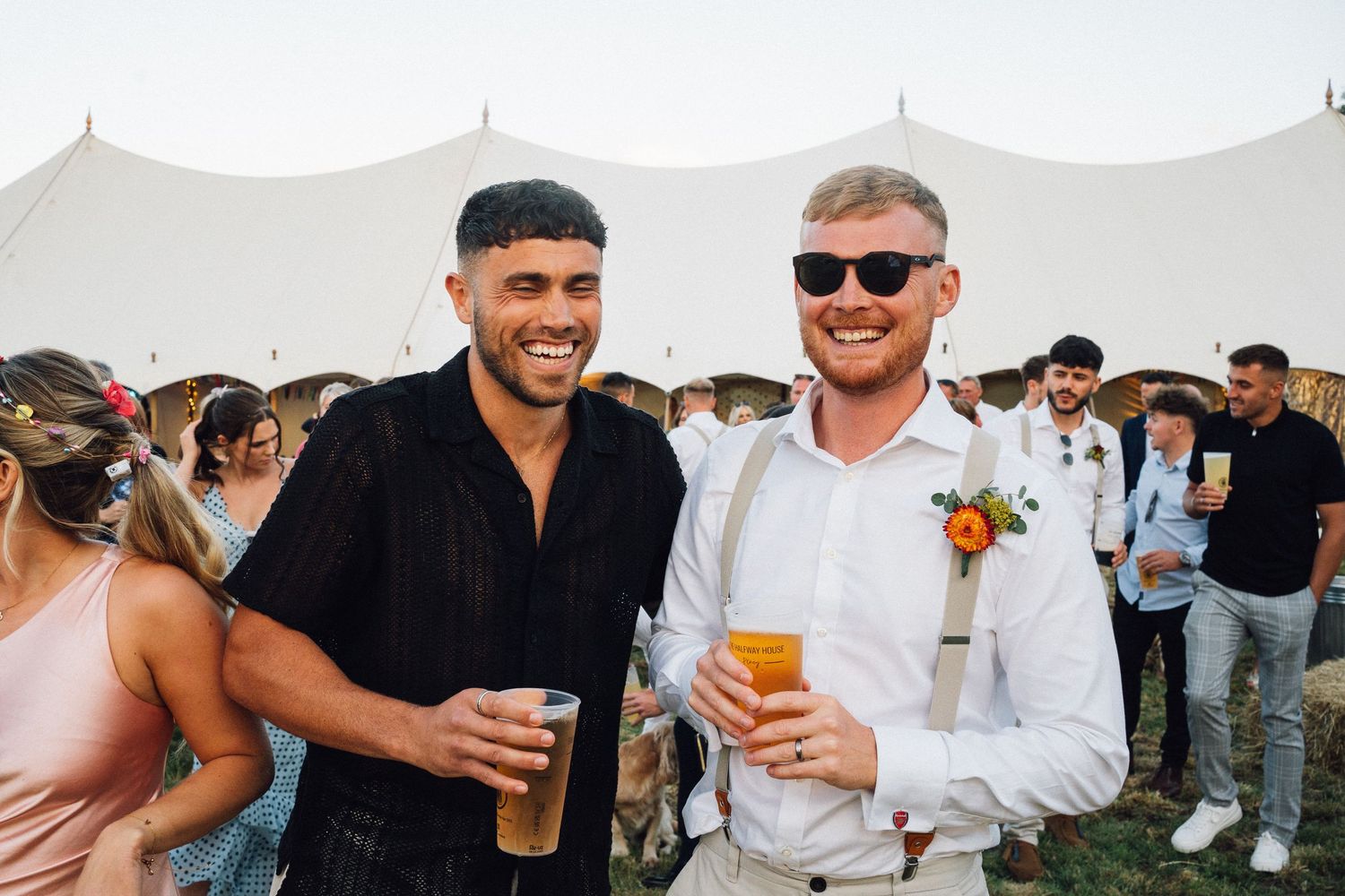 Wedding guests enjoying drinks and mingling at an outdoor reception under a white tent.