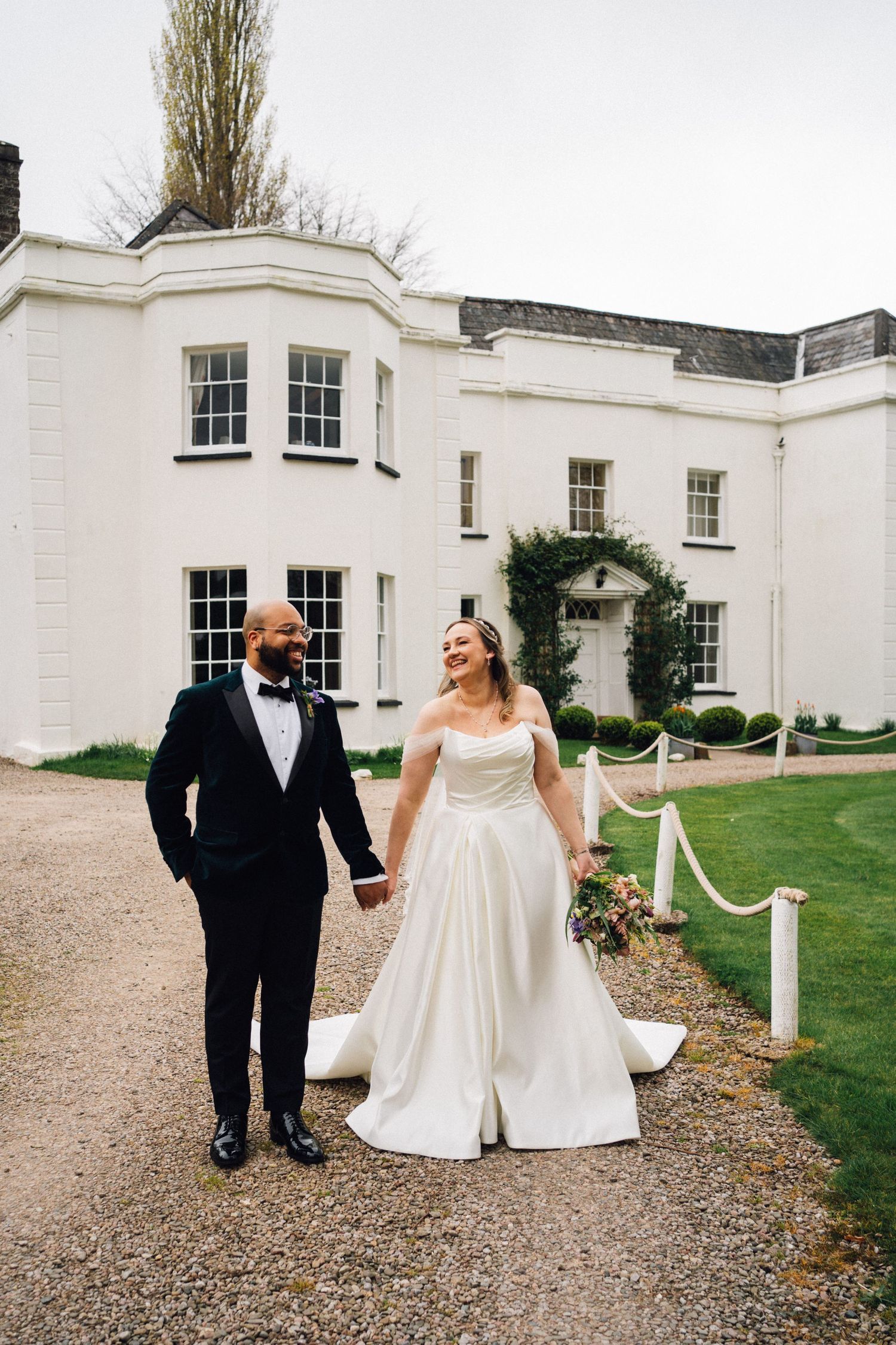 A newlywed couple walks hand in hand outside a grand white manor house on their wedding day.