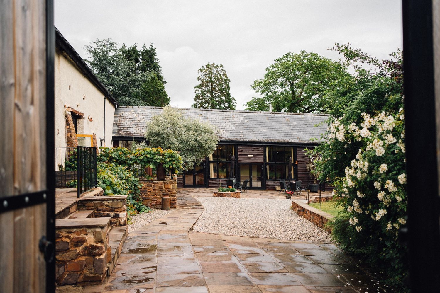Stone walkway leads through lush garden with blooming roses and rustic wooden pergola at luxury estate venue.