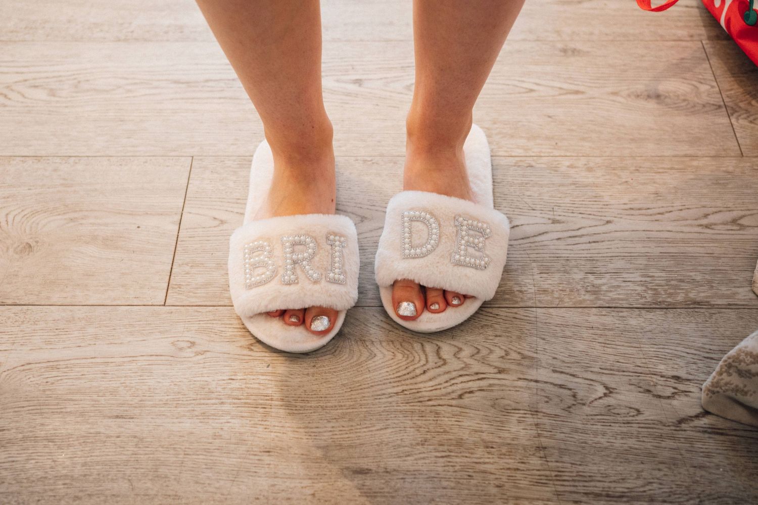 White plush slippers with 'BRIDE' text worn on wooden floor during wedding preparations.
