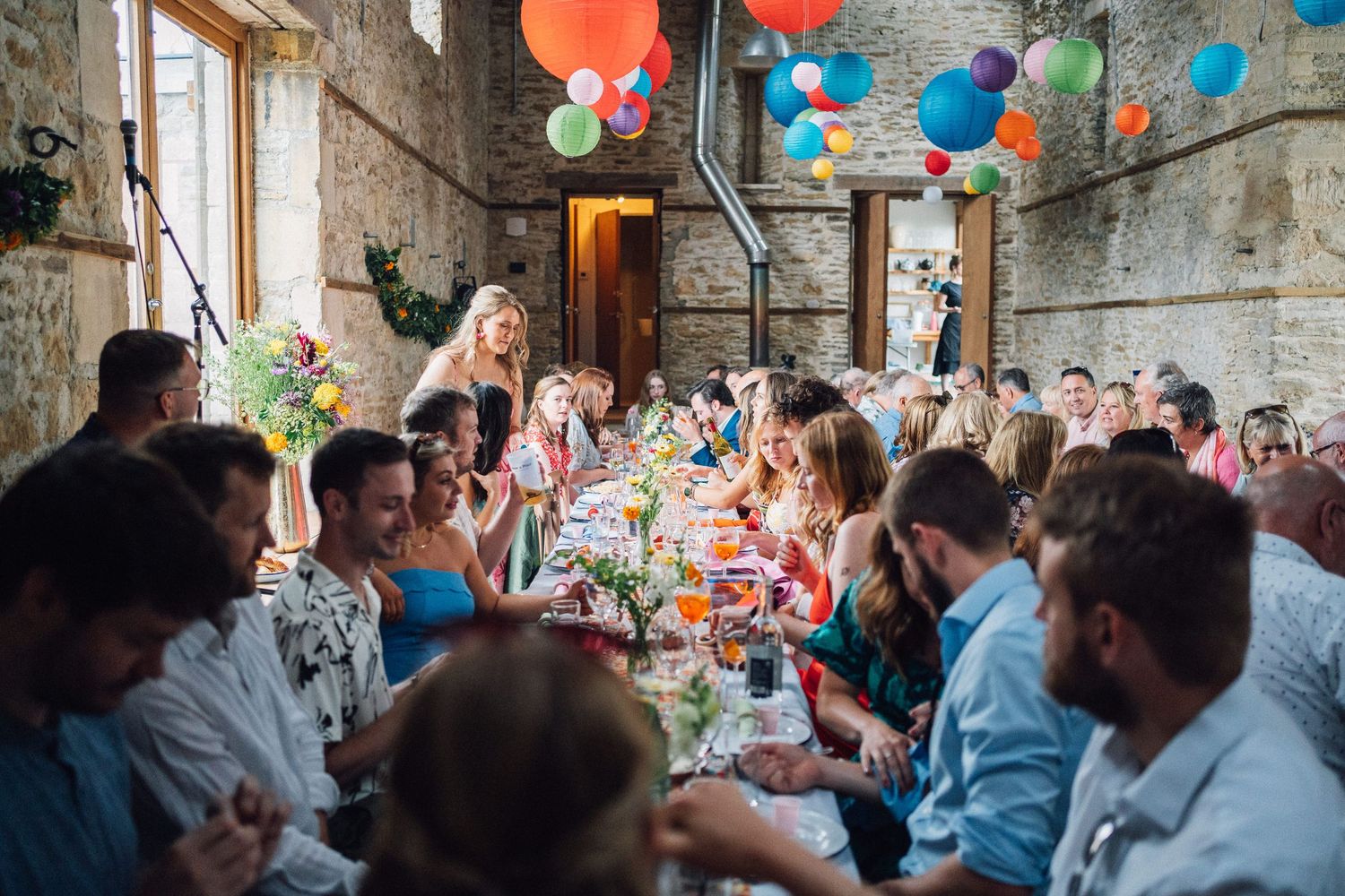 Guests enjoy a festive celebration at a rustic venue decorated with colorful balloons and string lights.