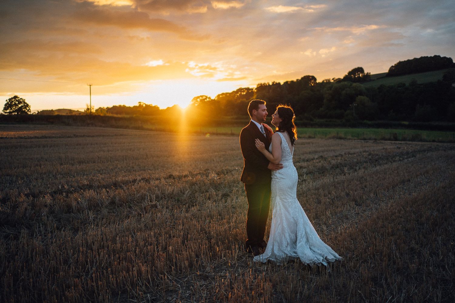 A couple stands silhouetted against a dramatic golden sunset in a harvested field as the sun dips below the horizon.