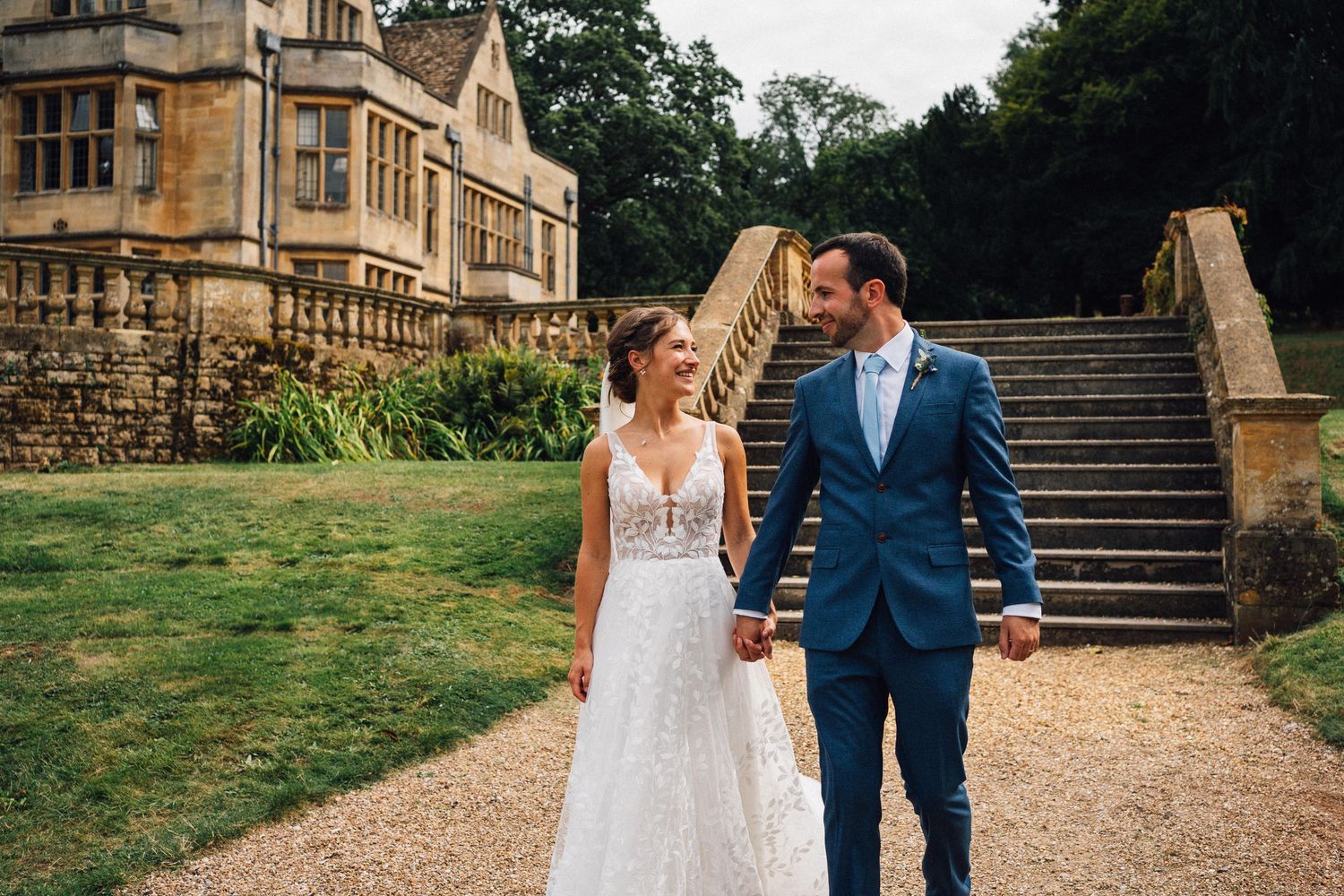 A couple walks hand in hand along a gravel path with grand stone steps and manor house behind them.