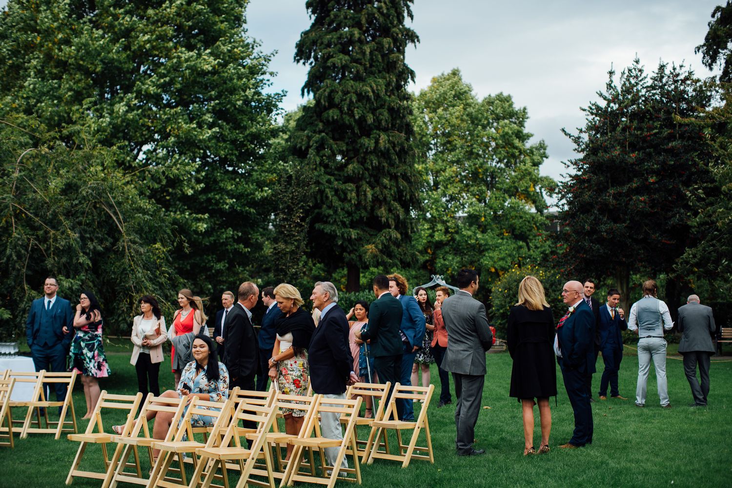 Guests gather on a lawn with wooden folding chairs set up for an outdoor ceremony among tall evergreen trees.