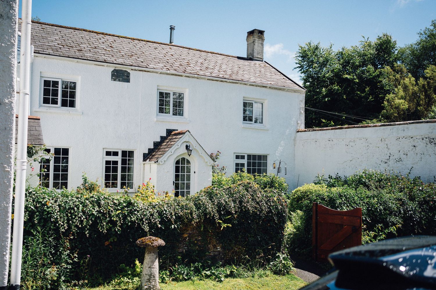 White two-story farmhouse with traditional architecture and lush garden border against a sunny backdrop.