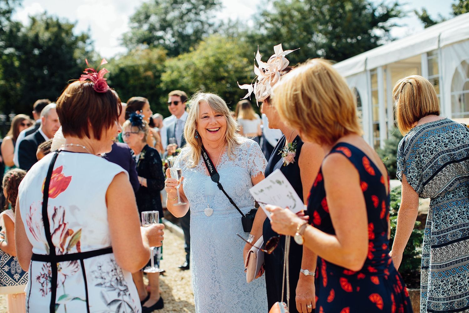 Wedding guests mingling outdoors on a sunny day with white tents in the background.