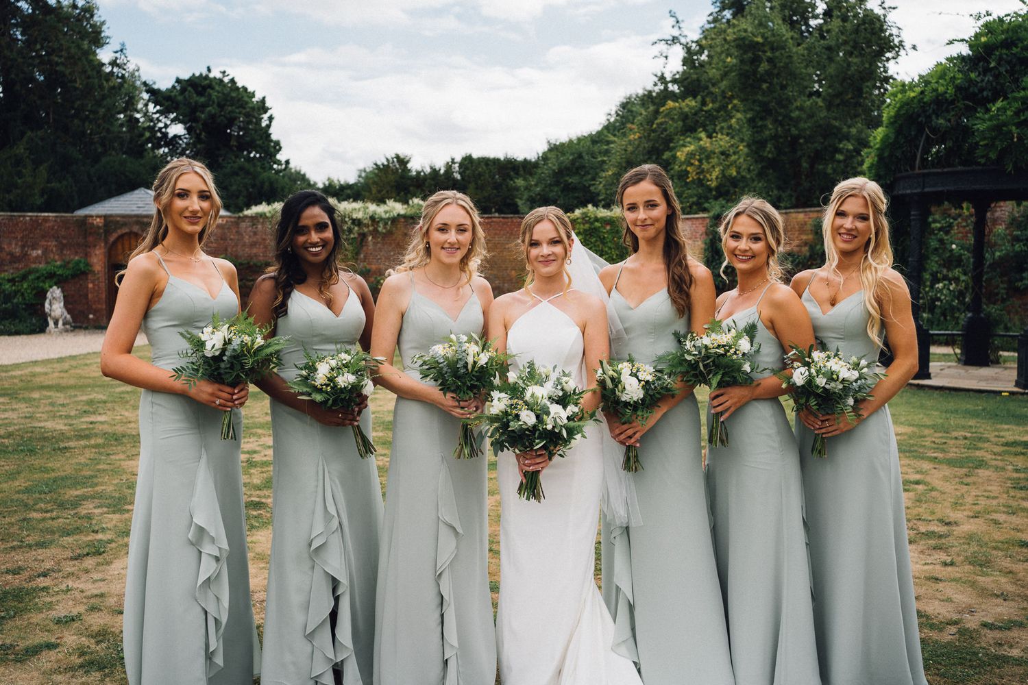 Wedding party of bridesmaids in pale gray dresses holding bouquets on lawn.
