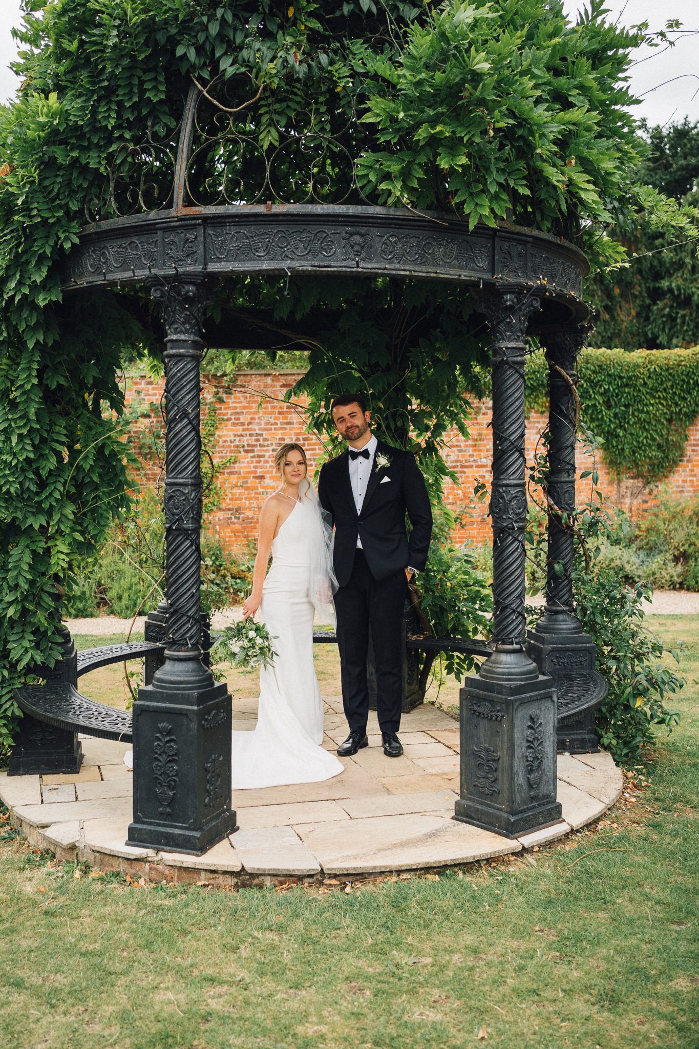 Elegant wedding couple poses under an ornate iron gazebo surrounded by green foliage.