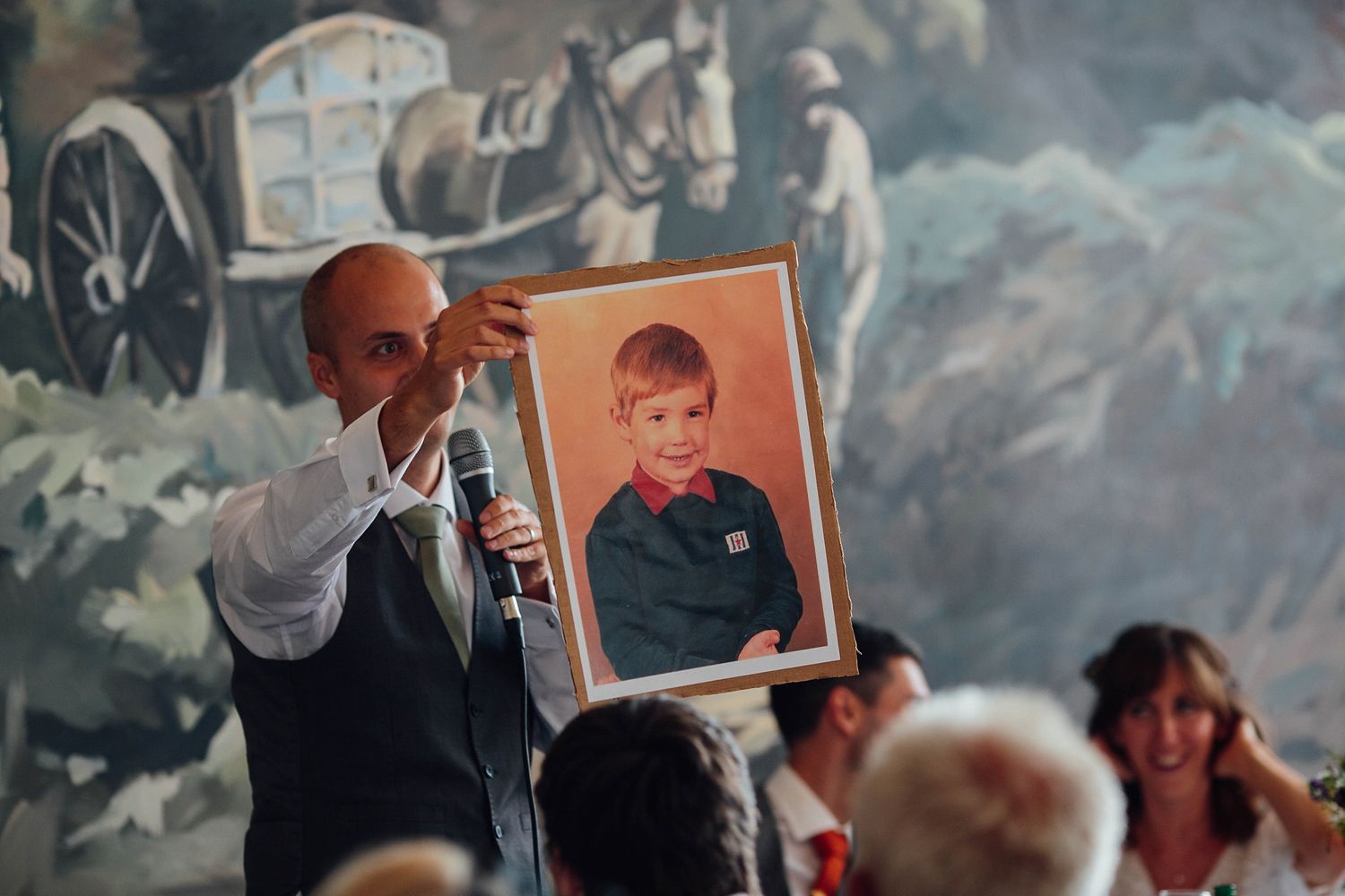 A speaker holds up a childhood photo during an emotional wedding speech against a painted mural backdrop.