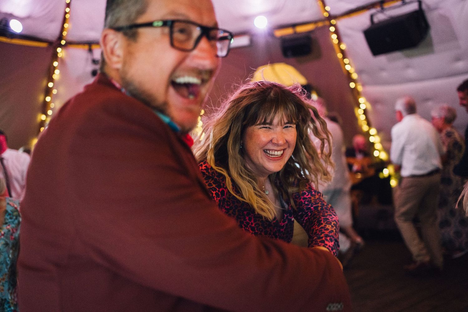 Lively wedding reception with guests dancing and celebrating under string lights and purple ambient lighting.