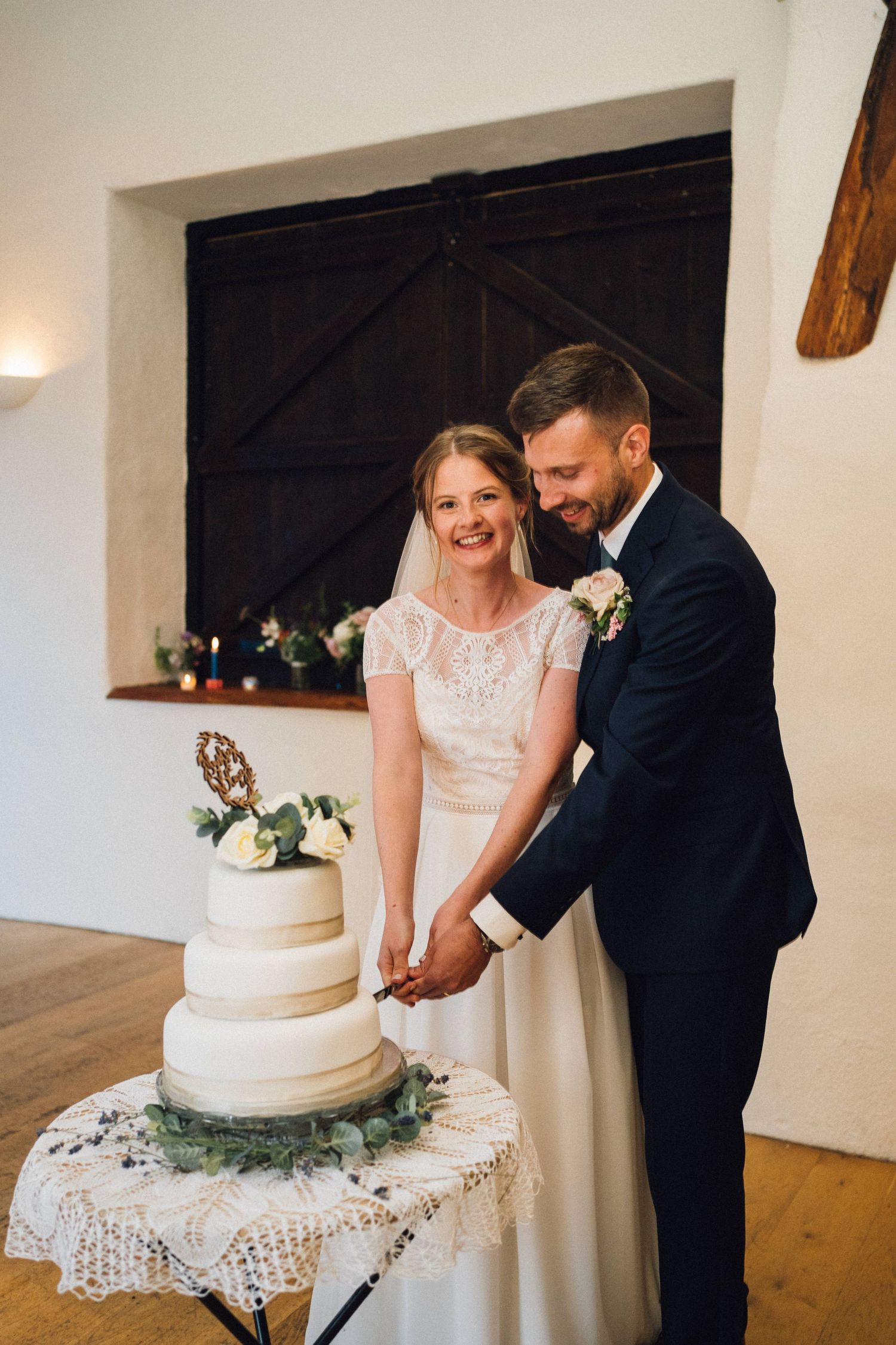 Couple cutting their white tiered wedding cake on a lace-covered table.