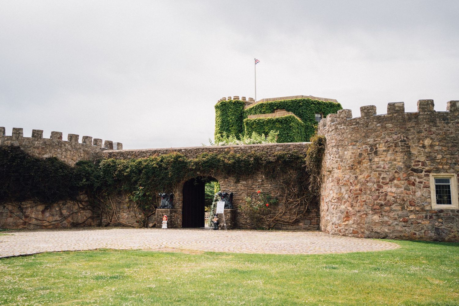 Historic stone castle entrance with ivy-covered battlements and visitors walking through arched gateway on cloudy day.