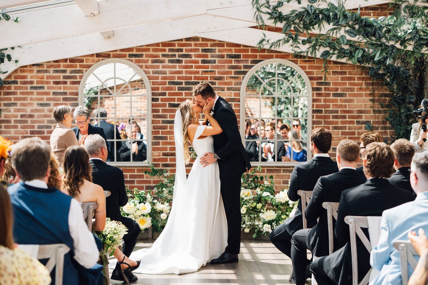Bride and groom share first kiss at elegant indoor wedding ceremony with brick walls and white floral arrangements.