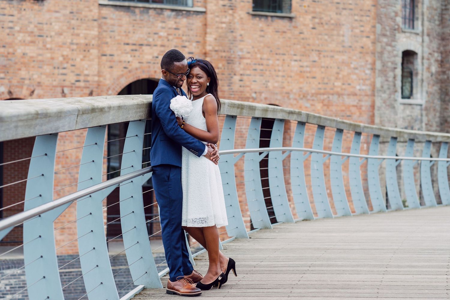 A couple shares a romantic moment on a curved bridge walkway with brick buildings in the background.