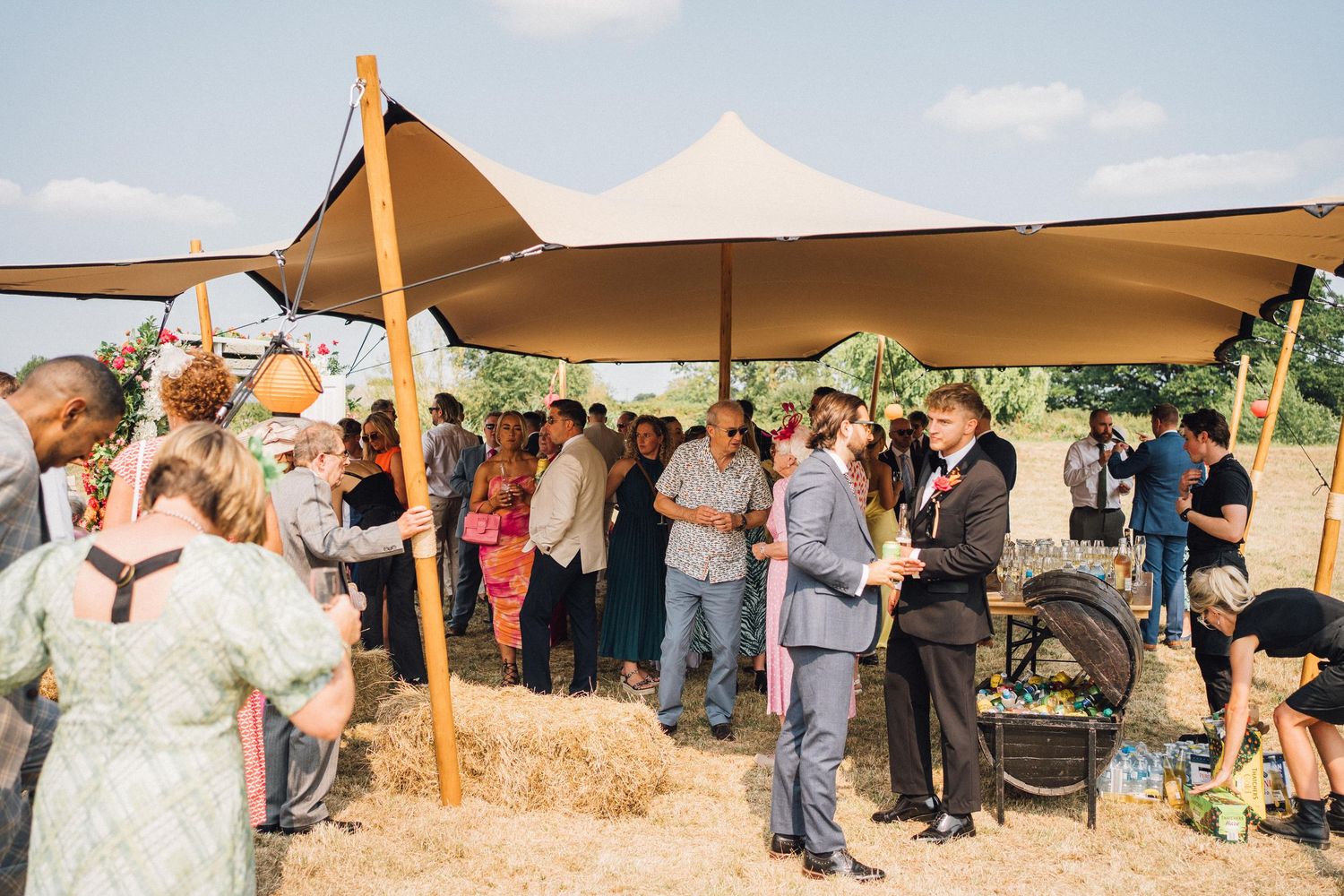 People gather under beige canopies at an outdoor event with hay bales and rustic decor.