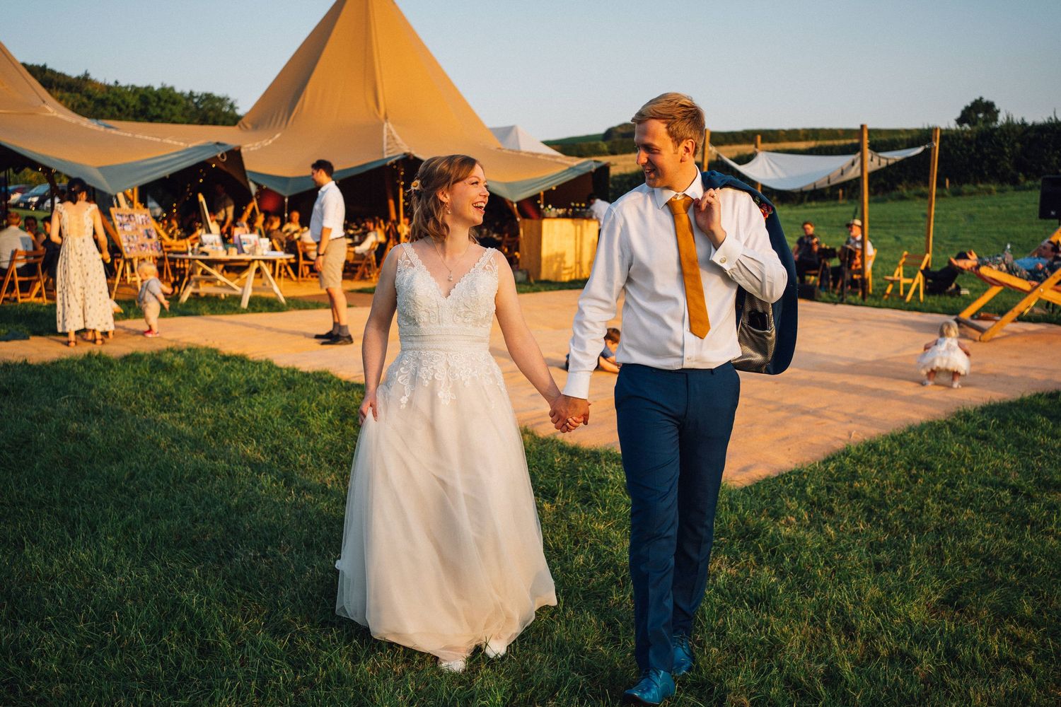 Wedding couple walks hand in hand at sunset near rustic teepee tents set up in a grassy field.