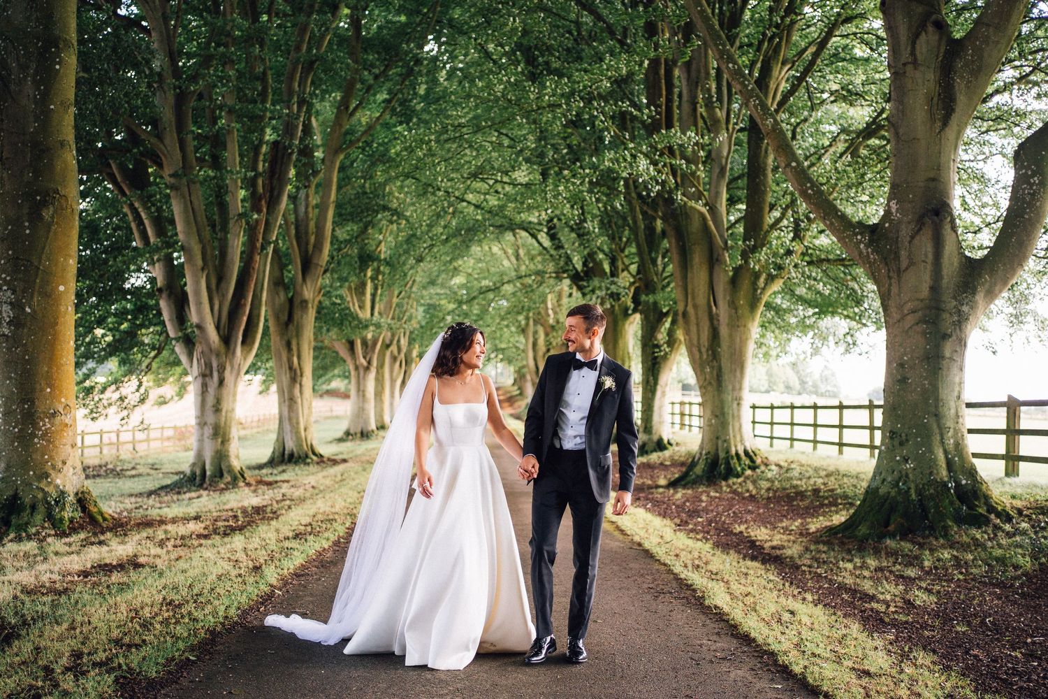 Newlywed couple walking down a tree-lined path with sunlight filtering through the branches.