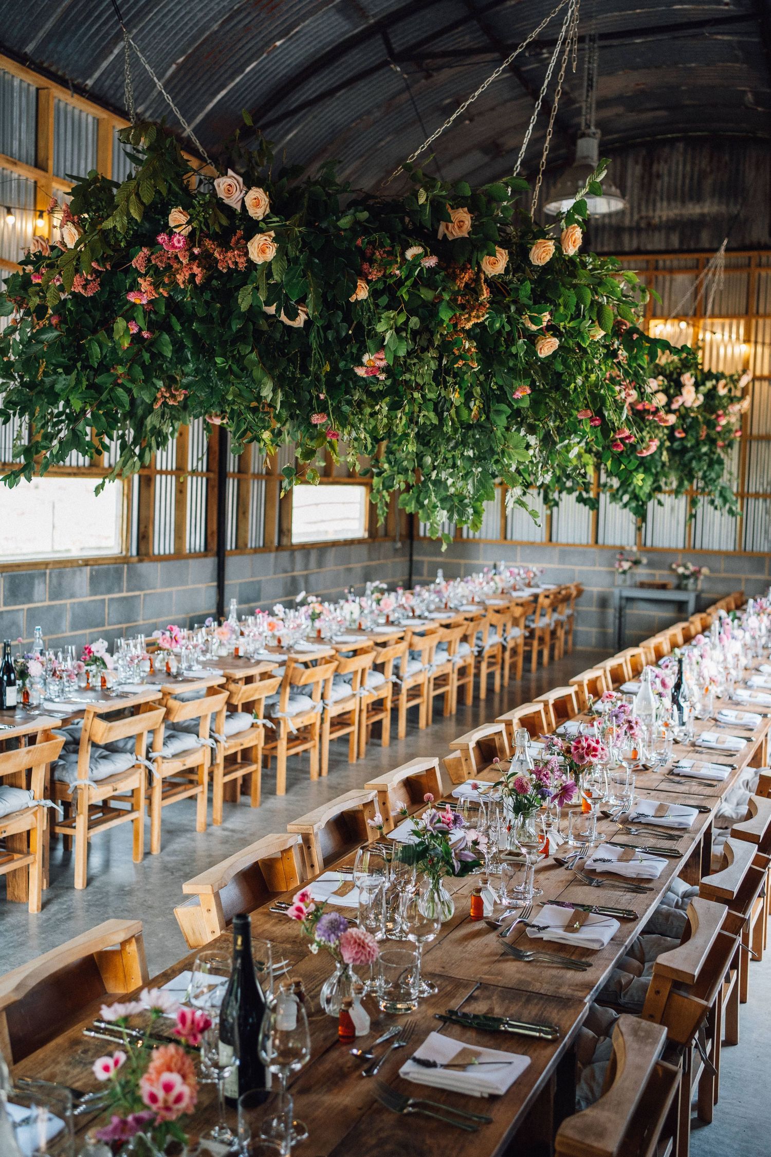 Long rustic dining tables set under hanging floral arrangements in an elegant barn wedding reception venue.