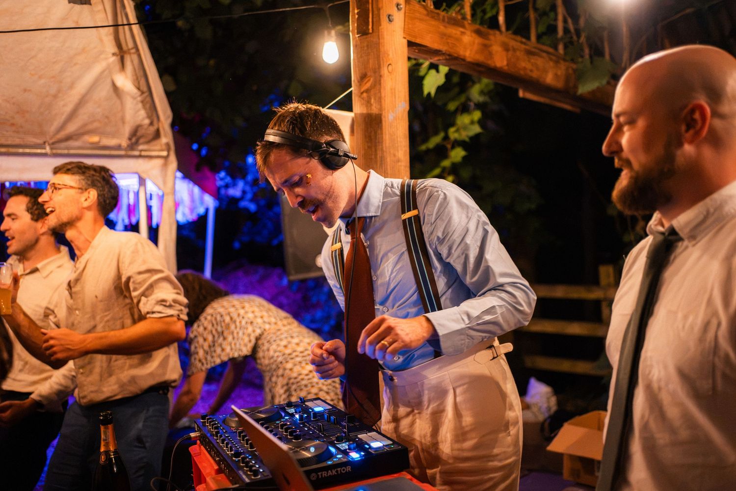 DJ wearing headphones operates mixing equipment at an outdoor evening wedding reception with string lights.