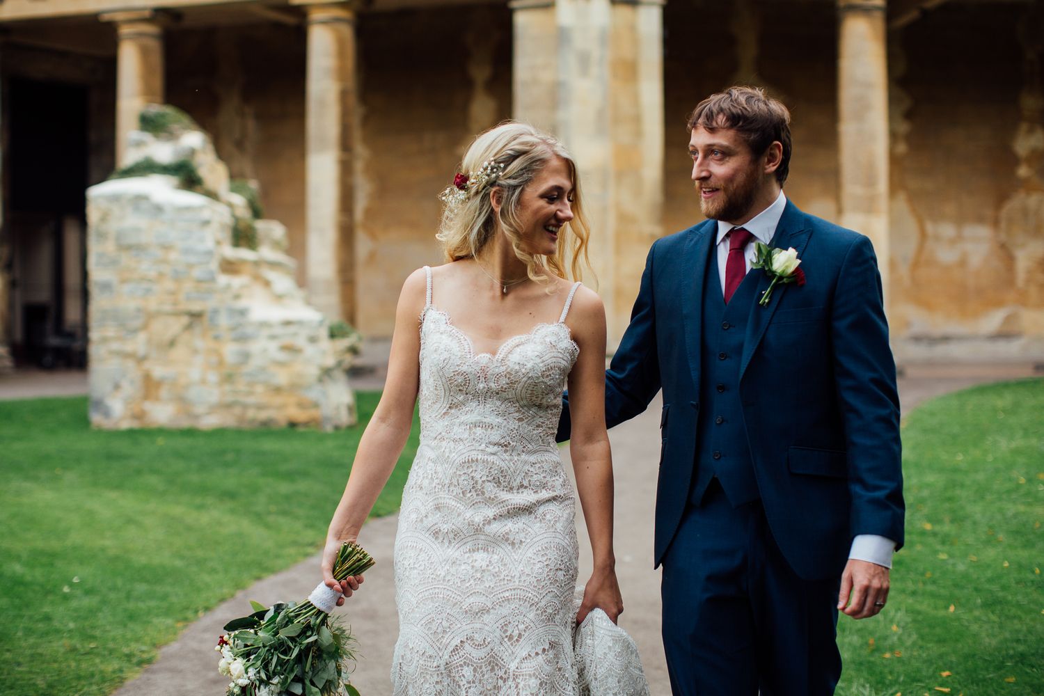 A couple in wedding attire walks together on a grassy lawn near a historic stone building.