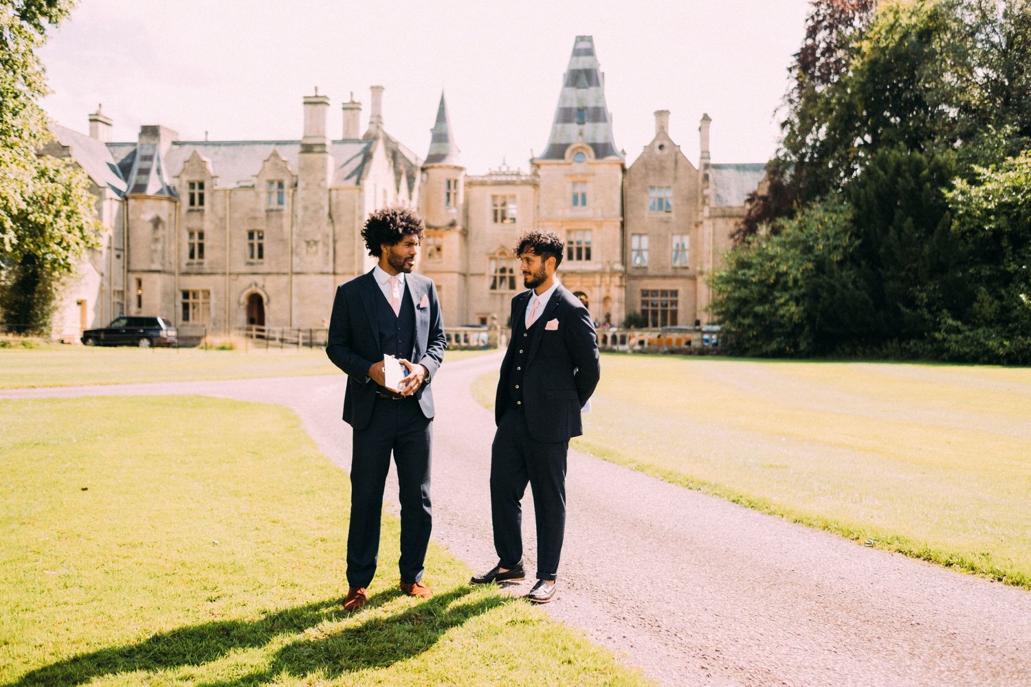 Two people in dark suits having a conversation on the lawn of a grand estate building.