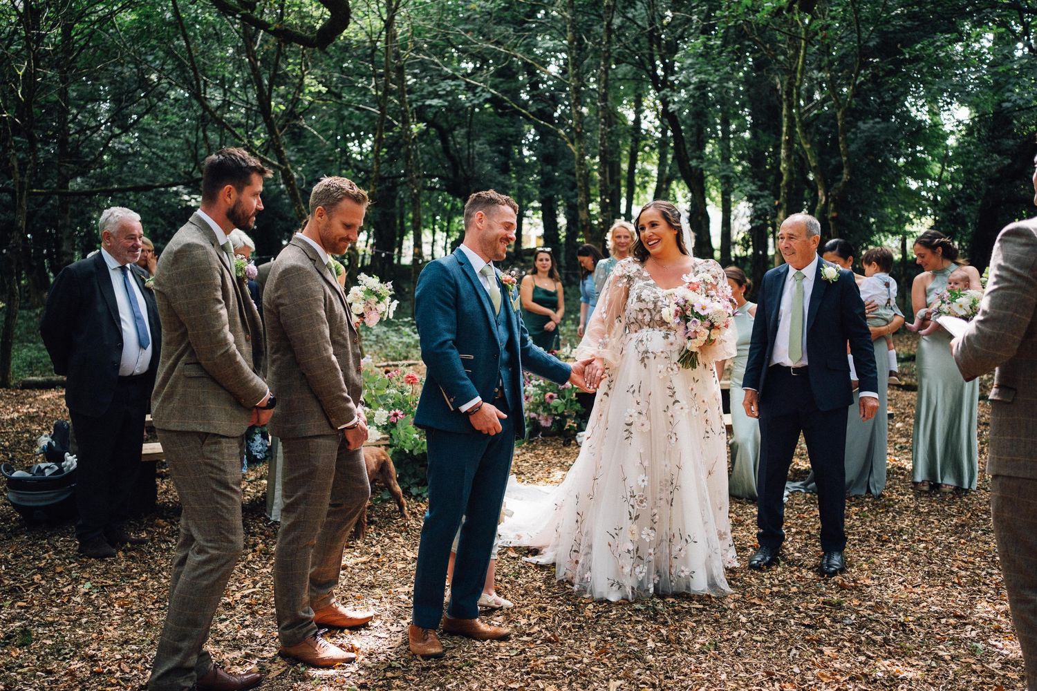 Wedding guests gather in a wooded outdoor ceremony space with natural lighting filtering through trees.
