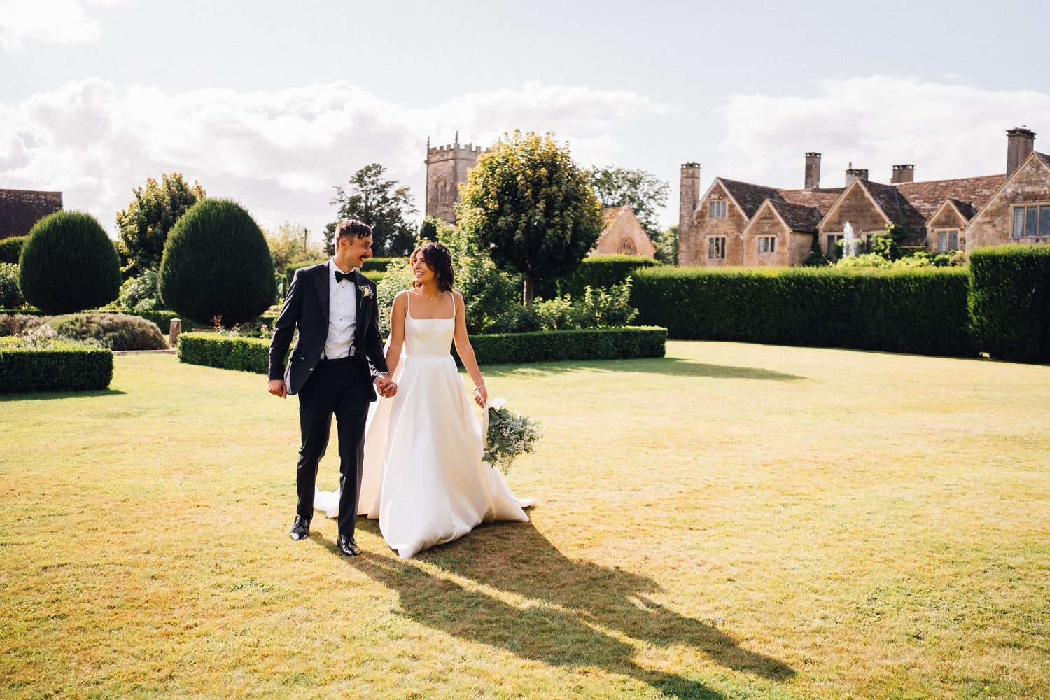 Newlyweds walking across a sunny lawn with historic buildings in the background.
