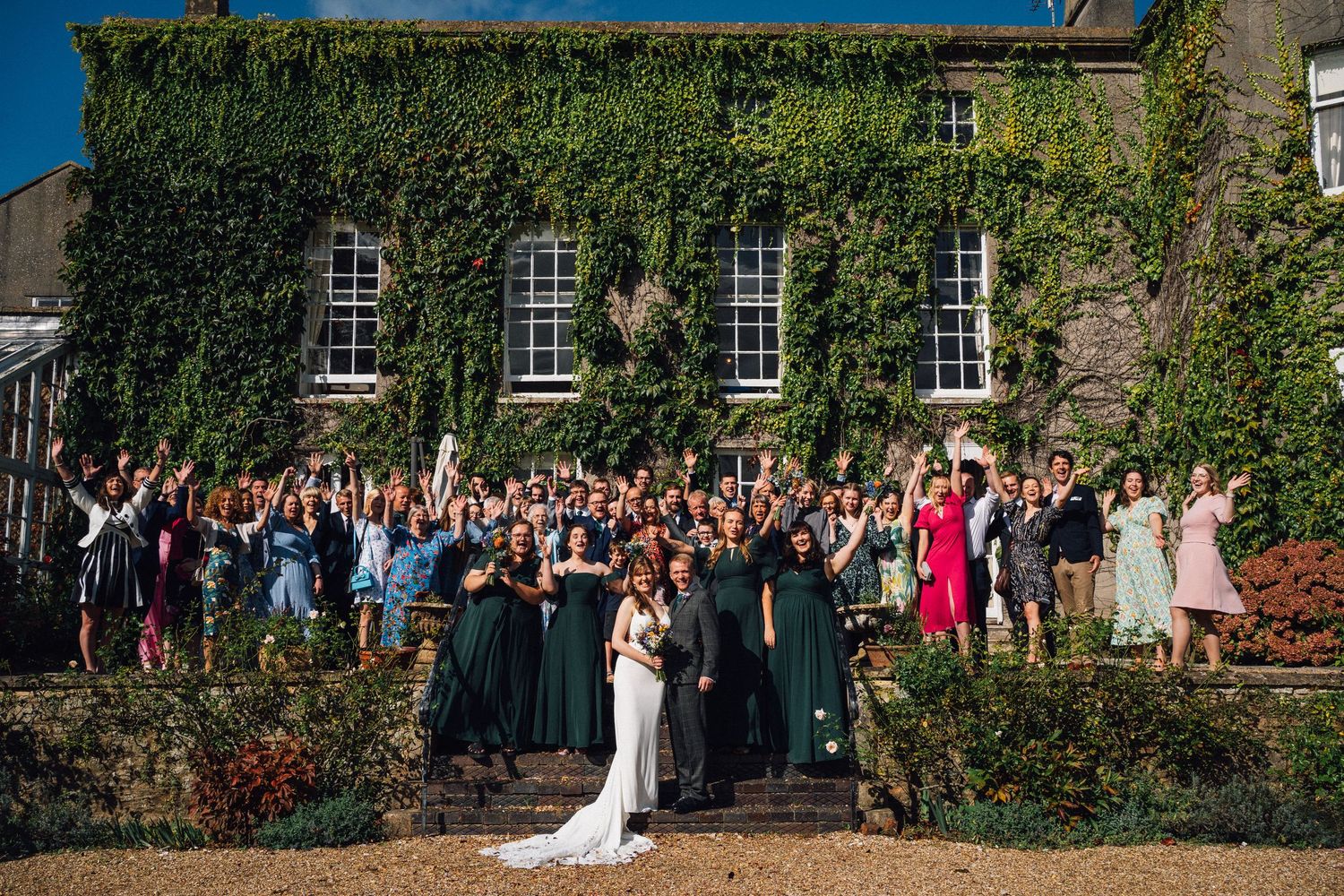 Large wedding group photo taken outside historic ivy-covered building.