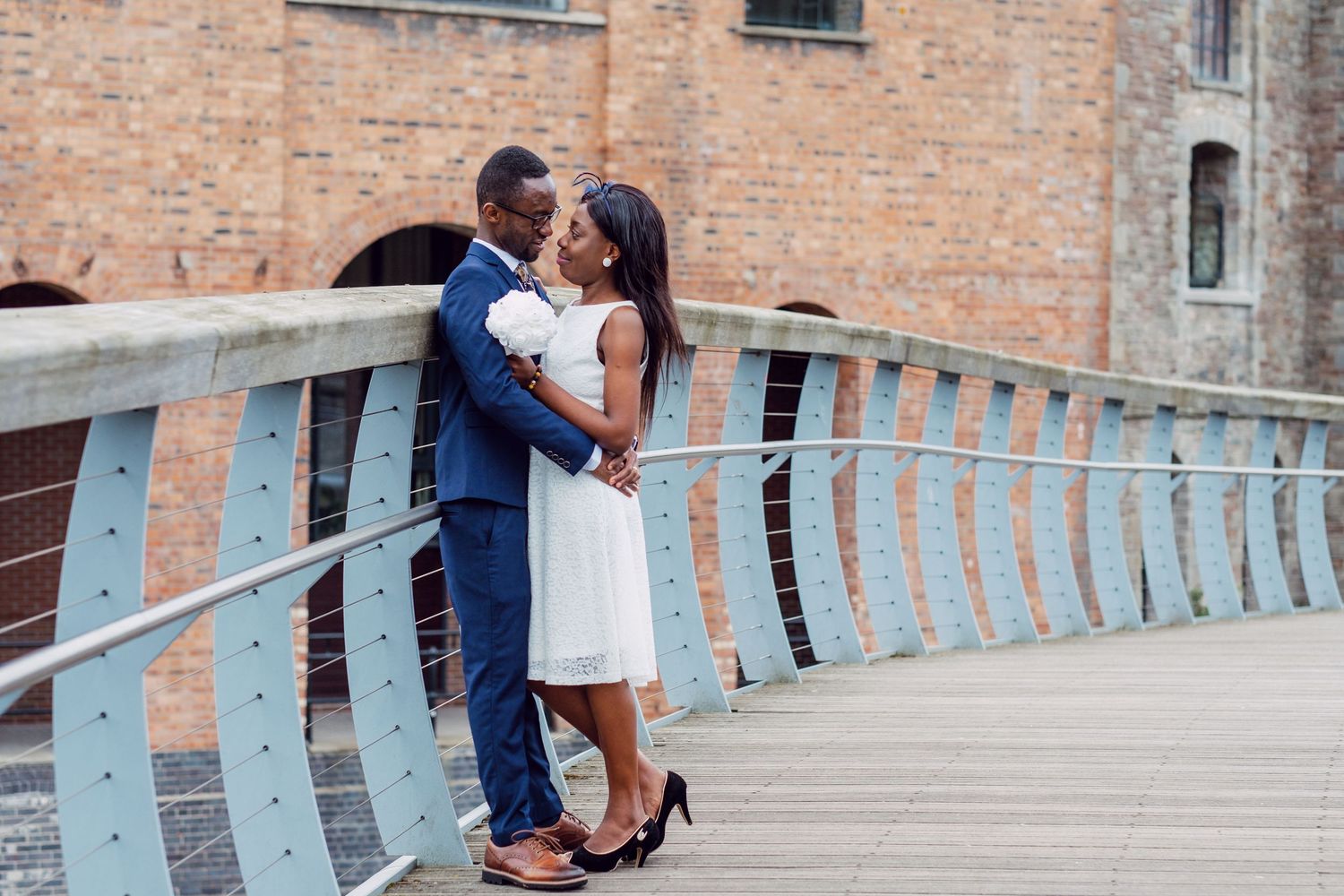 Romantic couple embracing on a bridge with blue railings and brick building backdrop.