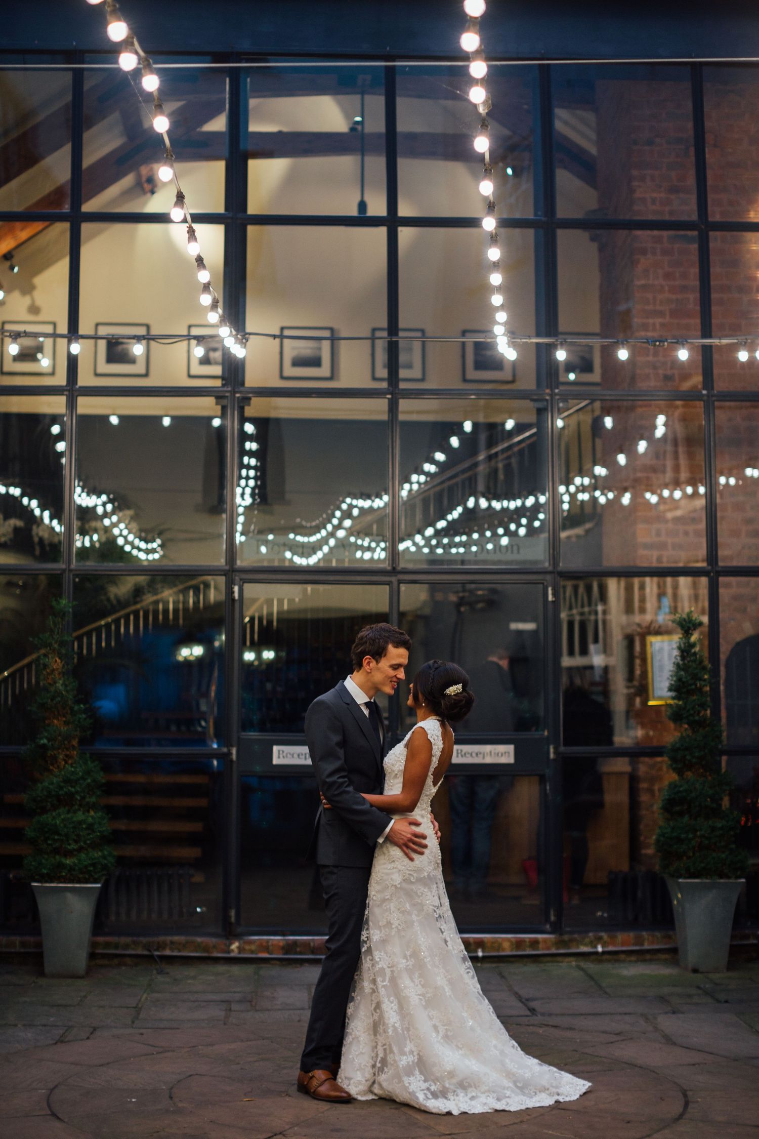 A romantic wedding couple embraces under twinkling string lights outside a glass-fronted venue at dusk.