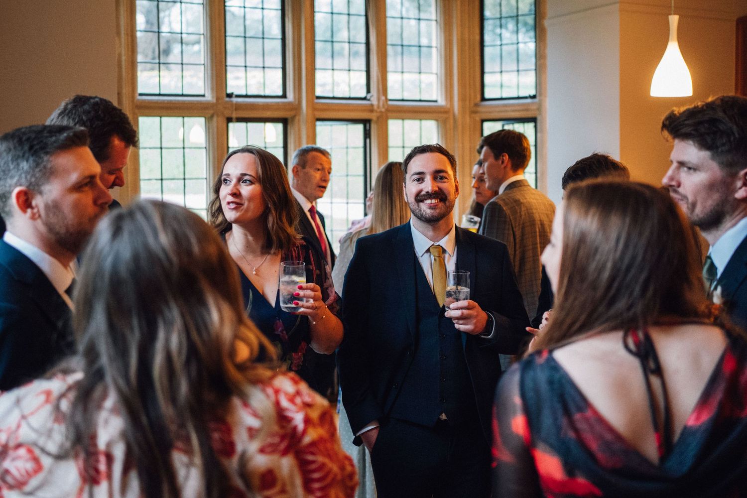 Wedding guests mingle and socialize in a bright indoor venue with large windows.