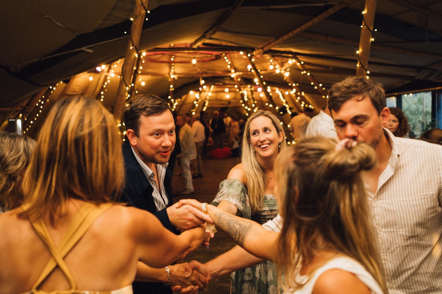 A group of people dancing and celebrating at a rustic barn wedding reception with string lights overhead.