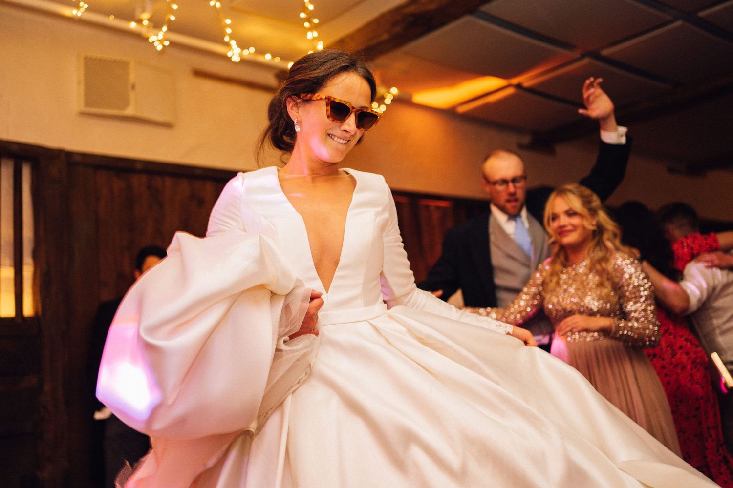 A person in a white wedding dress and sunglasses dances on the dance floor while guests watch in the background.