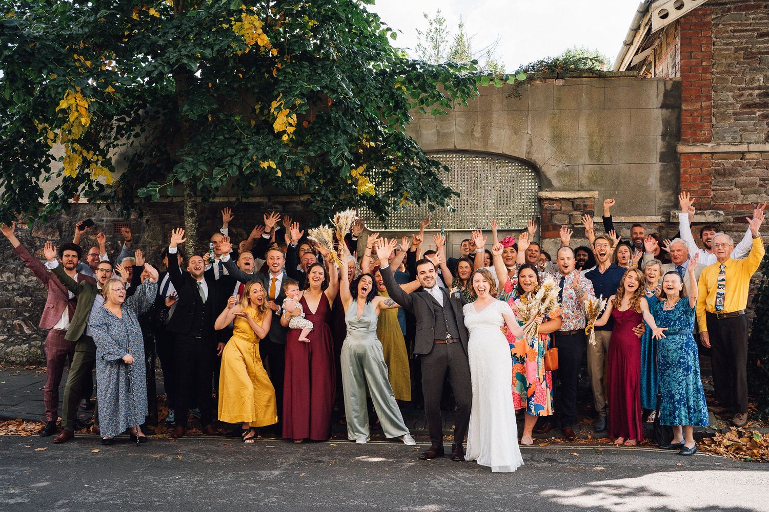 Wedding party members line up outside a brick building for a festive group photo in formal attire.