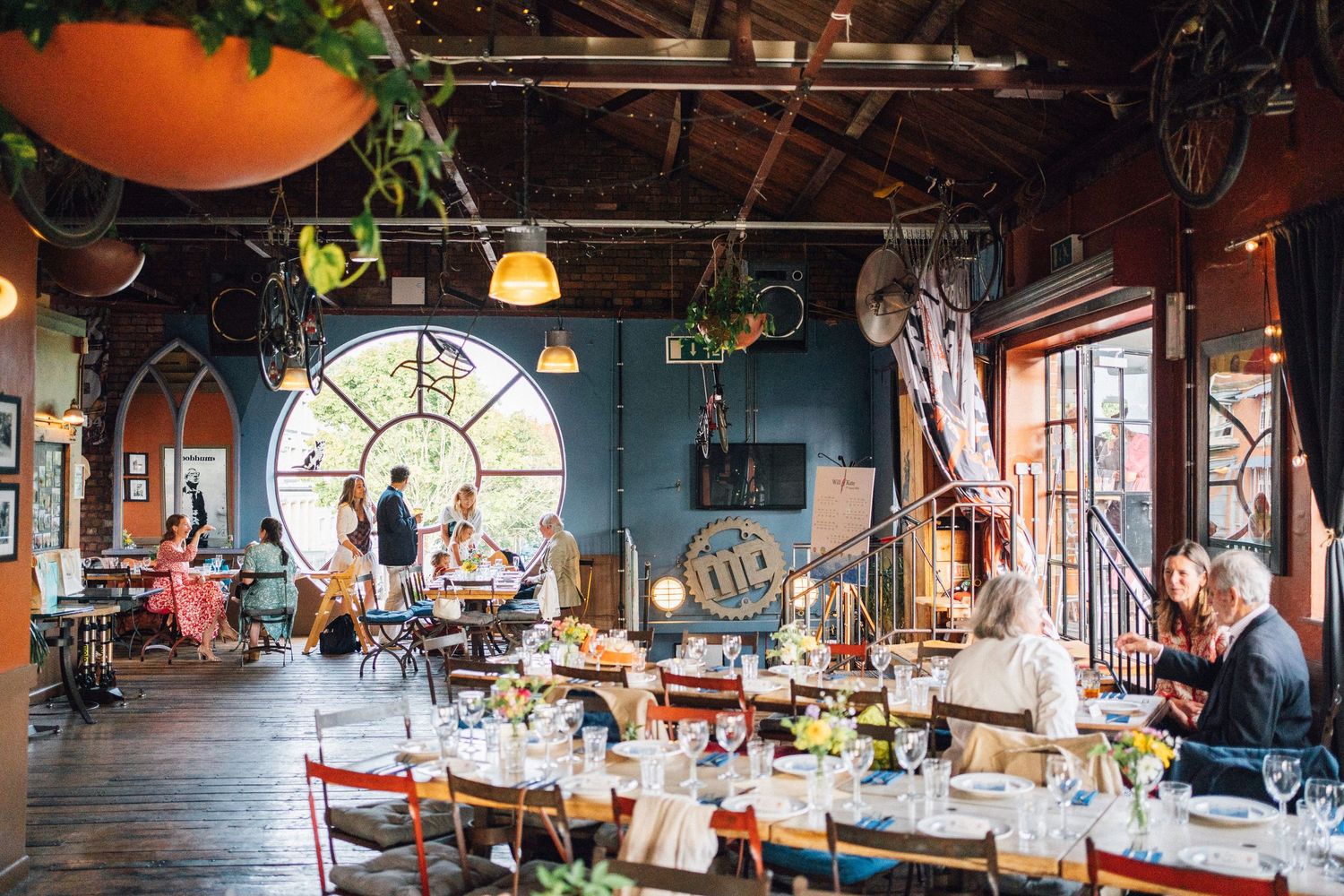 Eclectic restaurant interior with long dining tables, colorful chairs, round stained glass window, and lush hanging plants.