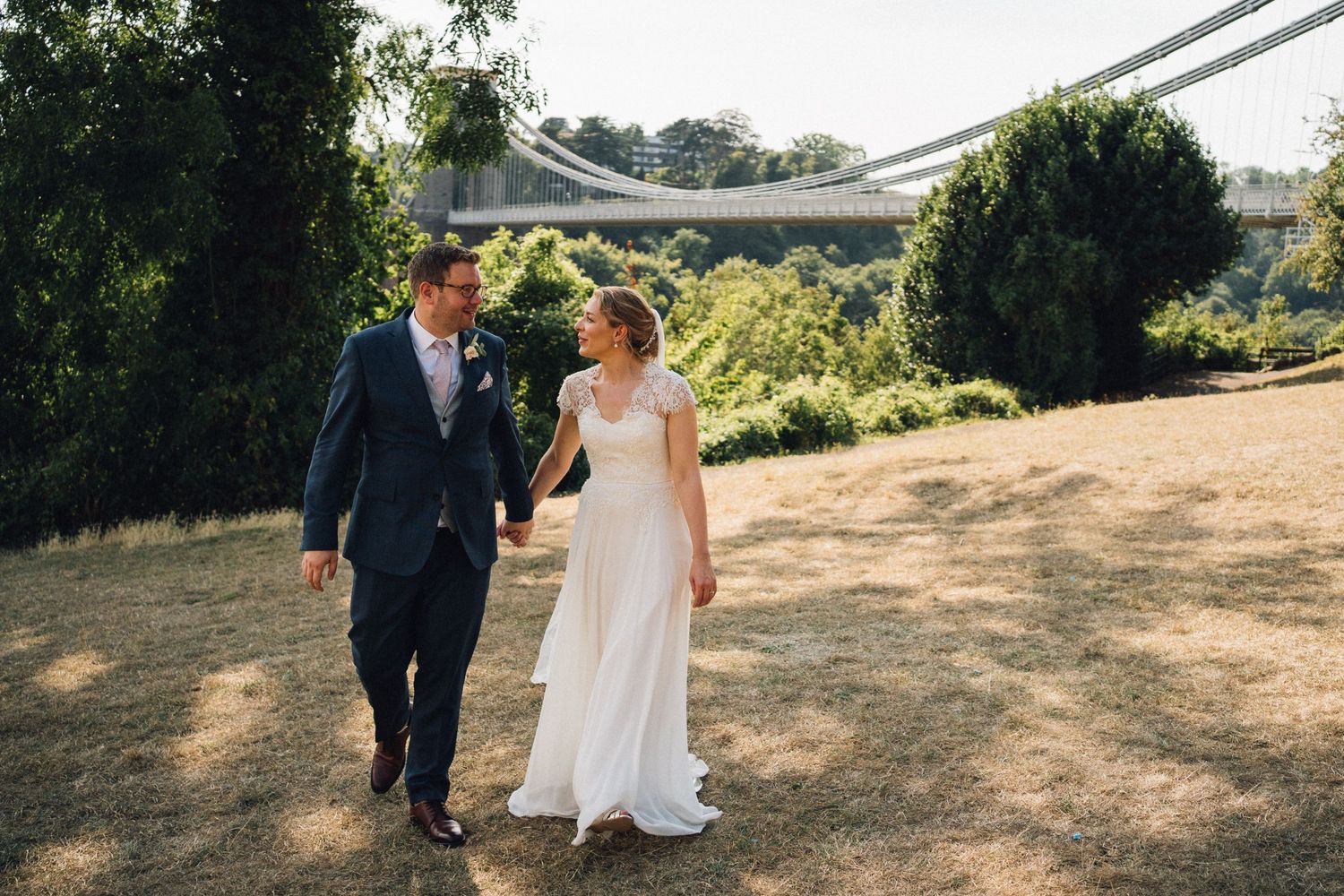 A bride and groom walk hand-in-hand on a sunny lawn with the Clifton Suspension Bridge visible in the background.