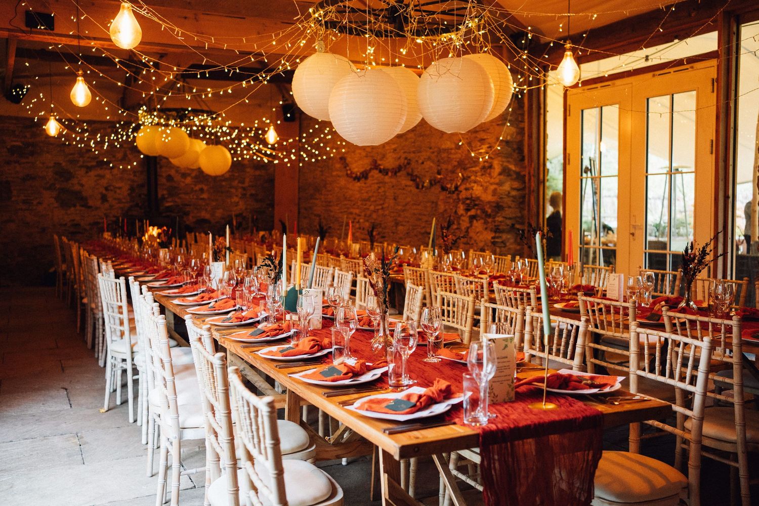 A rustic barn wedding reception venue decorated with string lights, white balloons and tables set with red linens.