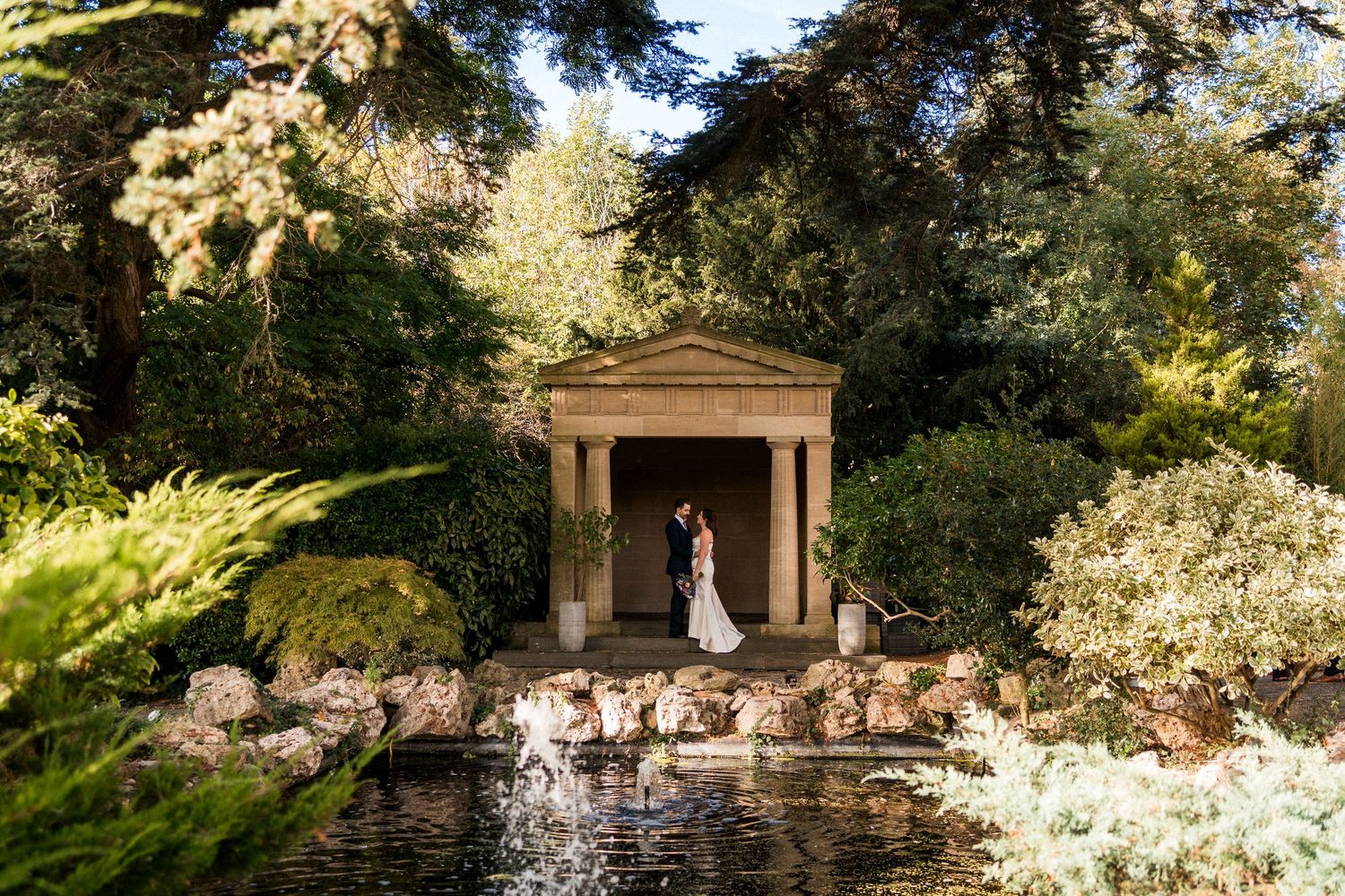 Bride and groom embrace inside a classical stone pavilion beside a tranquil pond with a fountain, surrounded by lush green garden foliage.