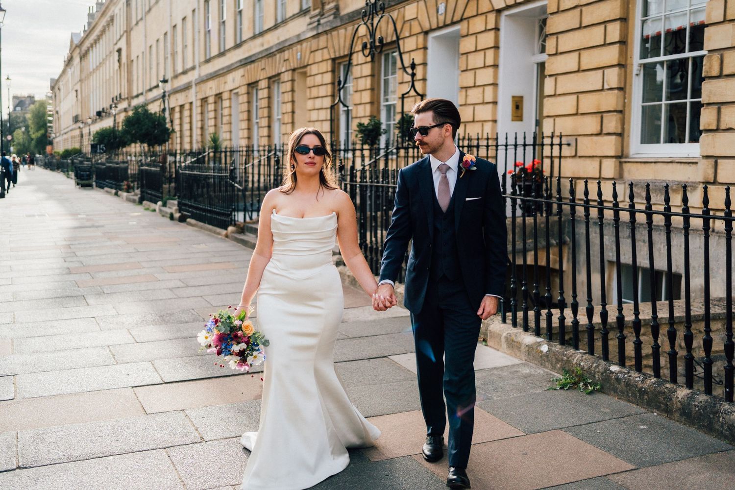 Bride in a white gown with a bouquet and groom in a navy suit hold hands while walking along a sunlit Georgian street.